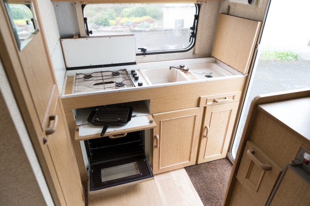 RV galley with timber cabinetry, sink, cooktop and open oven overlooking greenery